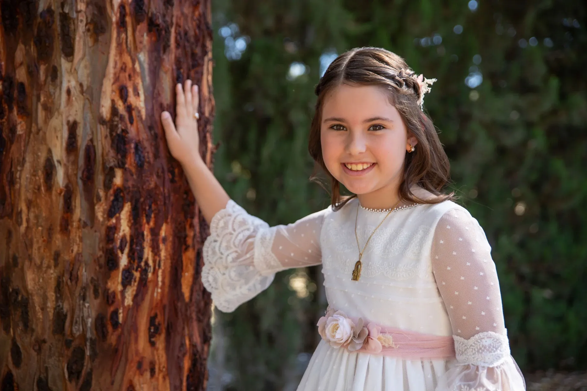 Niña de comunión sonriendo junto a un árbol